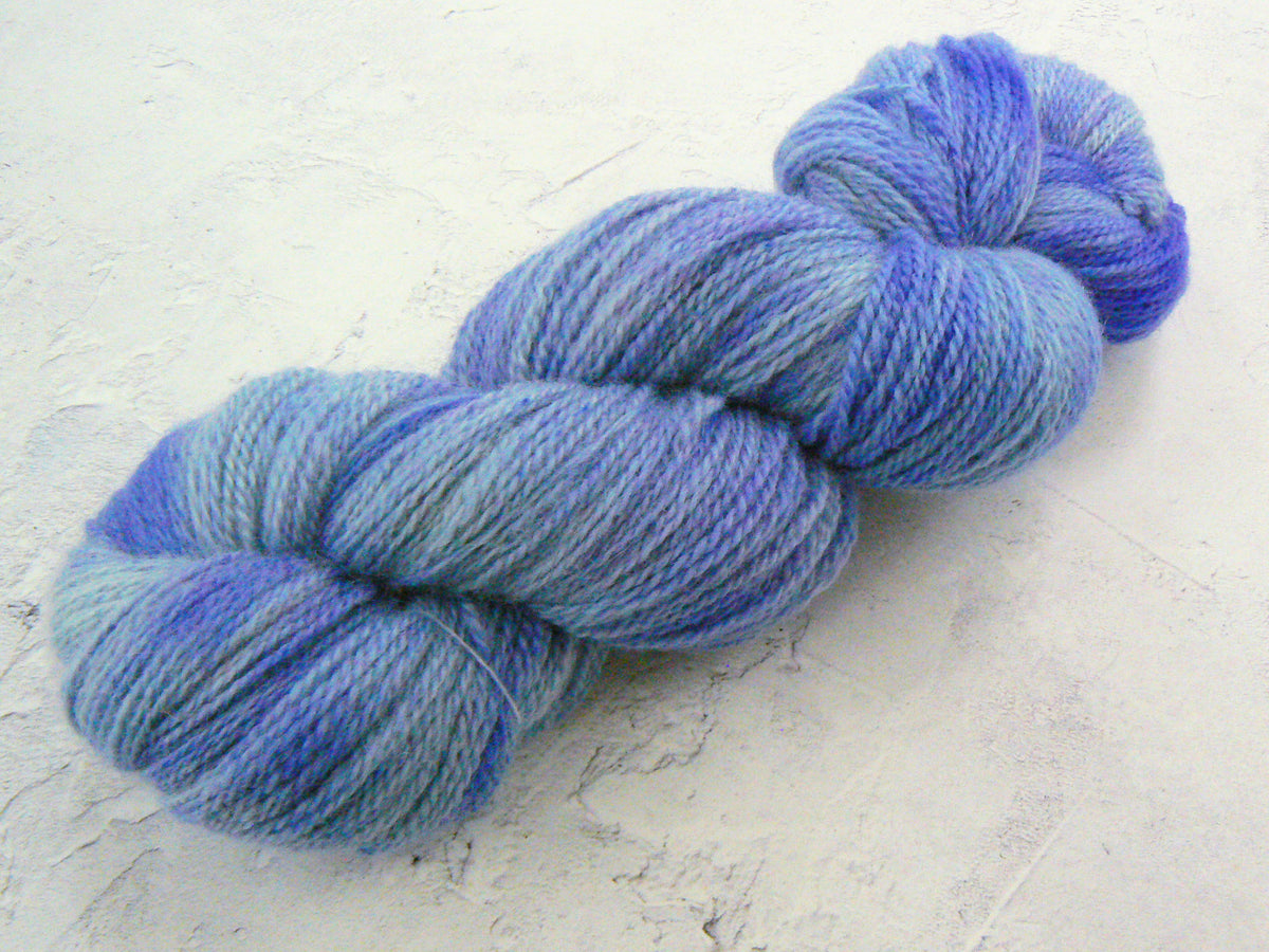 A twisted skein of variegated yarn in shades of blue and purple, resting on a textured off-white surface.