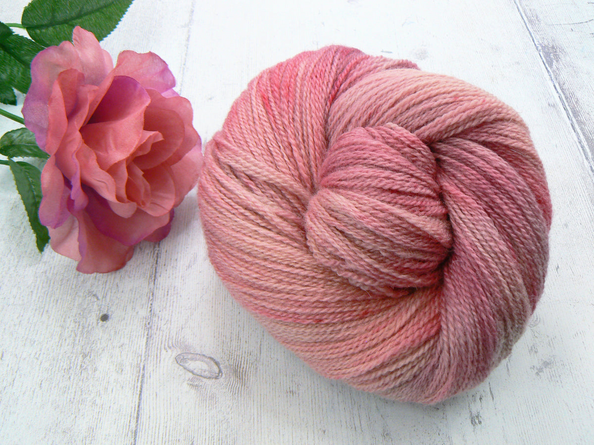 A skein of variegated pink and peach yarn next to a pink rose on a white wooden surface.