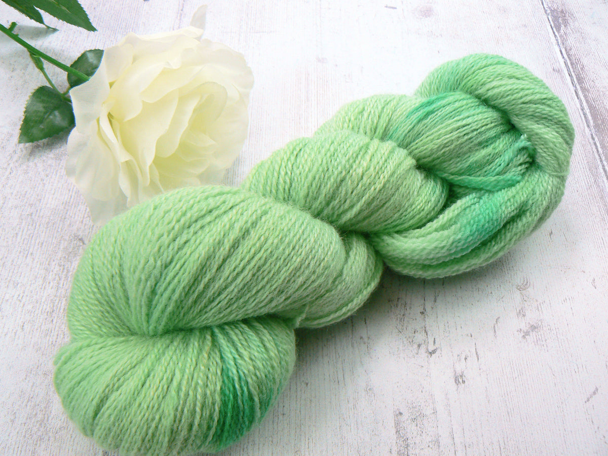 A skein of variegated mint green yarn arranged on a white wooden surface next to a white rose.