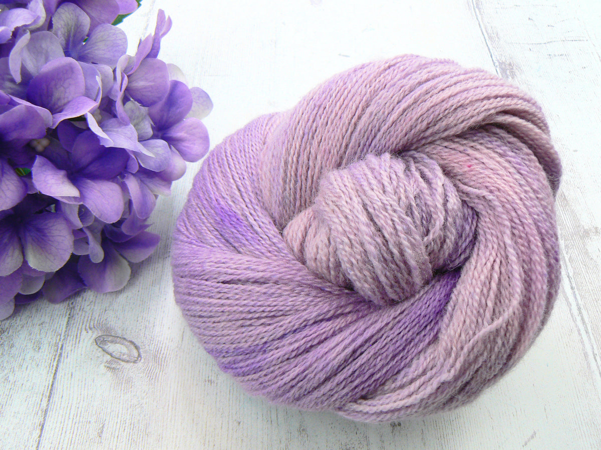 A skein of variegated lavender and purple yarn arranged in a knot on a white wooden surface, next to a purple hydrangea flower.