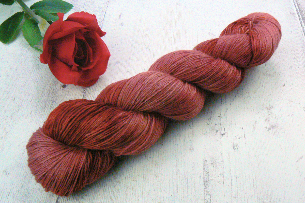 A twisted skein of dusty red-brown yarn displayed diagonally on a white wooden surface next to a red rose.