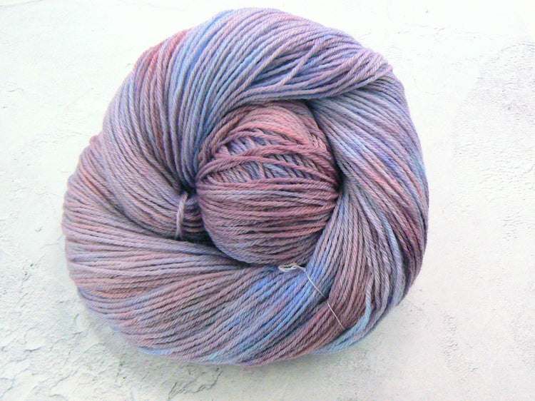 A skein of variegated yarn with blended lavender, pink, and light blue colors, arranged in a spiral shape on a textured off-white background.