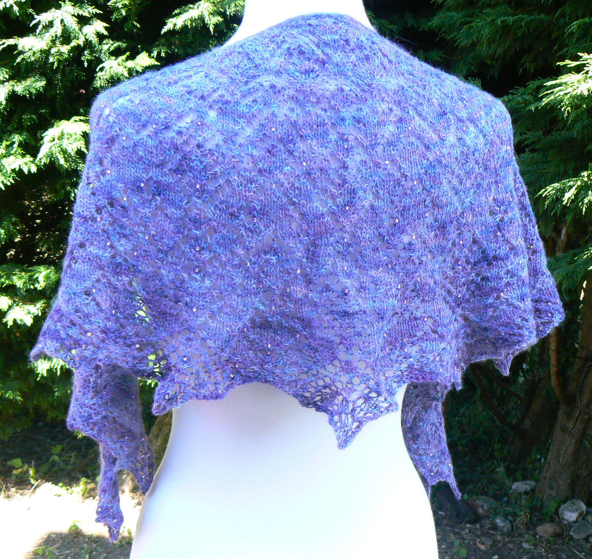A lavender purple open-knit shawl draped over a white mannequin torso, set against a background of green pine trees.