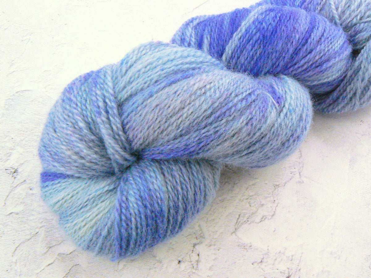 A skein of variegated yarn in shades of light blue, lavender, and medium blue, resting on a textured off-white surface.