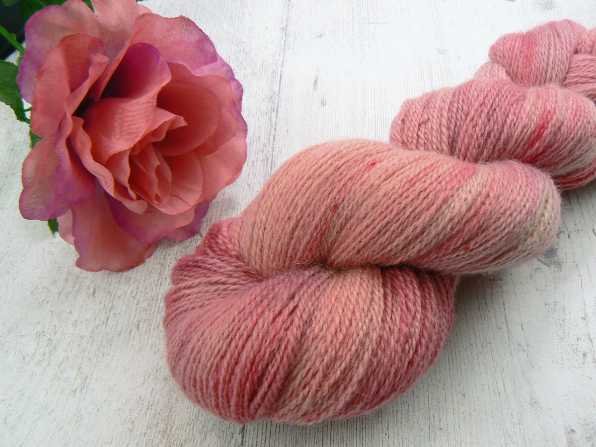 A twisted skein of variegated pink and peach yarn displayed on a white wooden surface next to a pink rose.