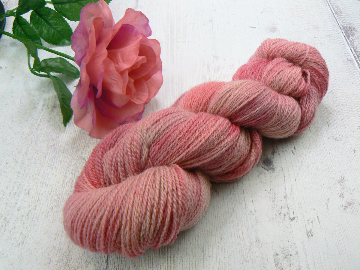 A twisted skein of variegated pink yarn displayed on a white wooden surface next to a pink rose.