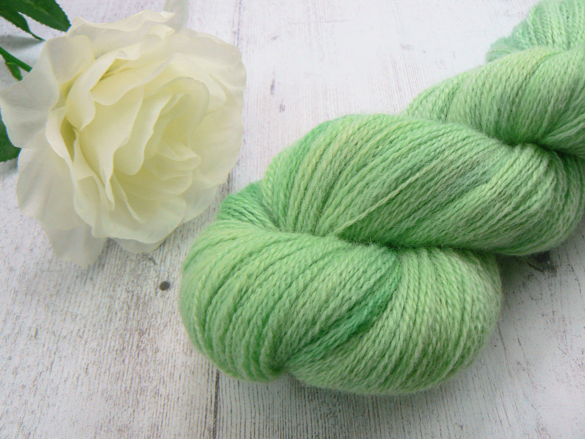 A skein of soft mint green yarn arranged on a white wooden surface next to a white rose.
