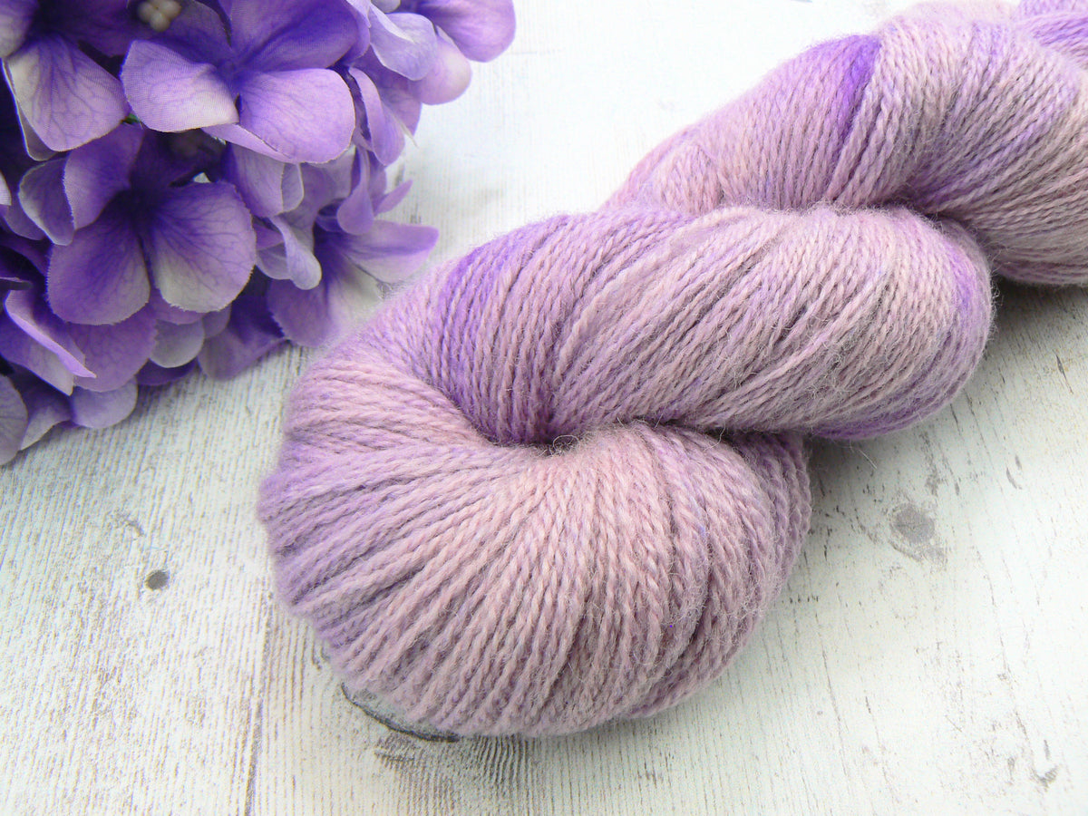 A twisted hank of soft lavender-purple yarn placed on a light weathered wood surface, with purple flowers partially visible in the upper left corner.