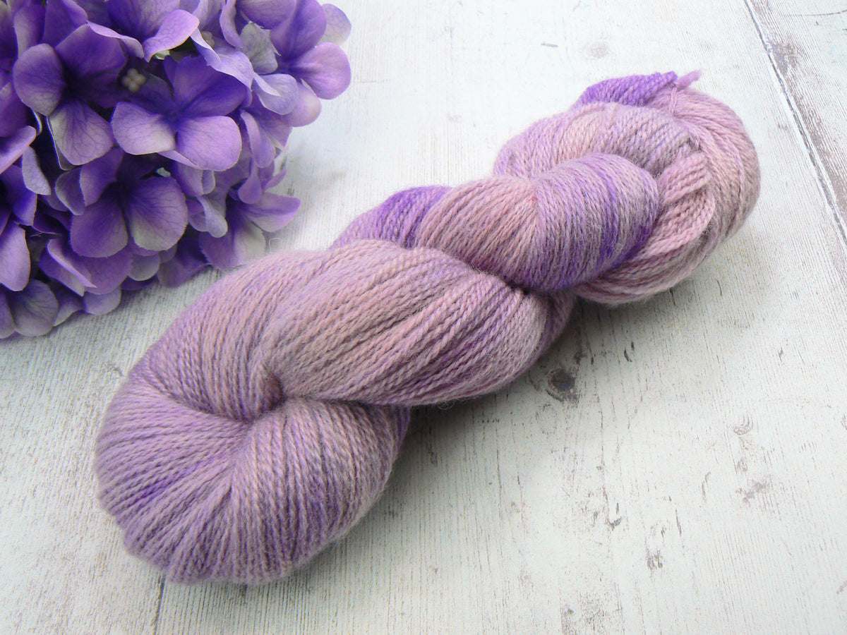 A twisted skein of variegated lavender and light pink yarn displayed on a white wooden surface next to purple flowers.
