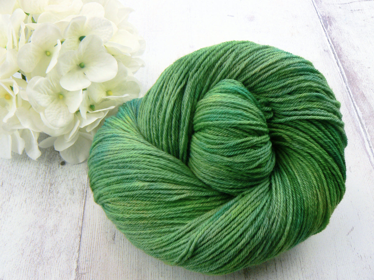 A skein of variegated green yarn arranged in a spiral on a light white wooden surface, styled next to white hydrangea flowers.