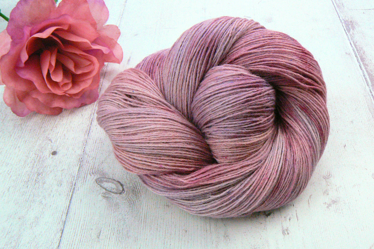 A skein of variegated yarn in dusty rose and mauve shades, styled on a white wooden surface next to a pink rose.