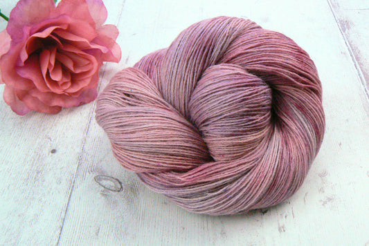 A skein of variegated yarn in dusty rose and mauve shades, styled on a white wooden surface next to a pink rose.
