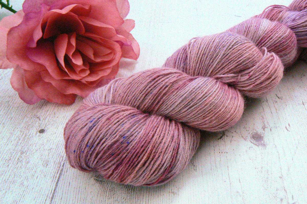 A hank of variegated pink and mauve yarn placed on a white wooden surface next to a pink rose.