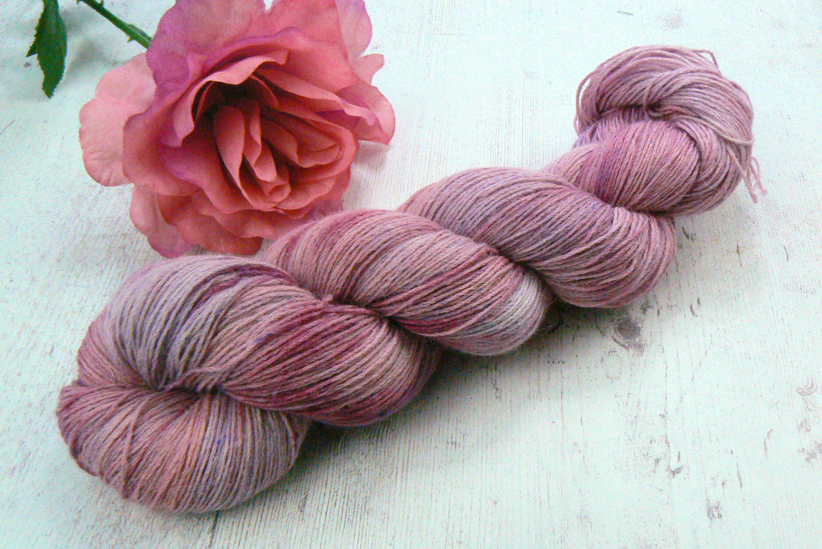 A twisted skein of variegated yarn in dusty mauve, pink, and lavender tones, styled with a pink rose on a white wooden surface.