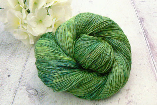 A skein of variegated green yarn arranged in a swirl on a light whitewashed wooden surface, with white flowers in the upper left corner.