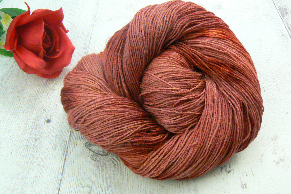 A skein of variegated yarn in a warm rust-brown colour, styled next to a red rose on a white wooden surface.