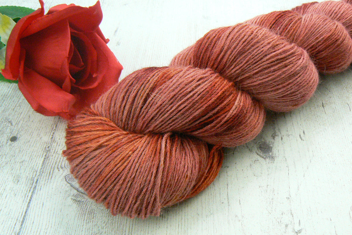 A skein of variegated yarn in warm rust, terracotta, and orange-brown tones, styled on a white wooden surface next to a red rose.