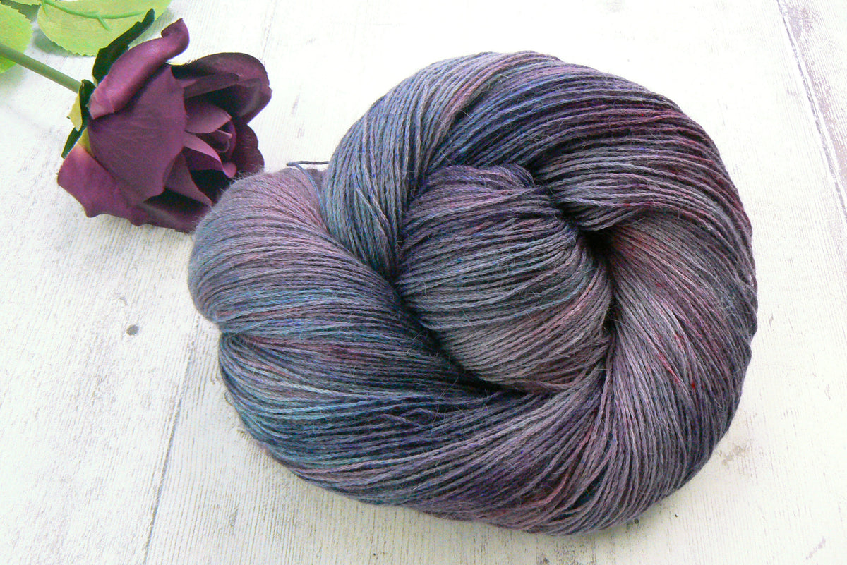 A skein of variegated yarn with blended purple, blue, and gray tones, styled on a white wooden surface next to a dark purple rose.