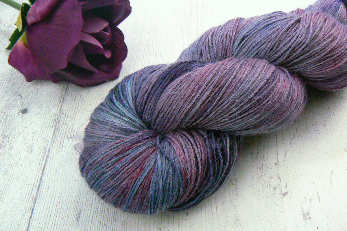 A twisted skein of variegated yarn with blended purple, blue, and pink tones, styled on a white wooden surface next to a dark purple rose.