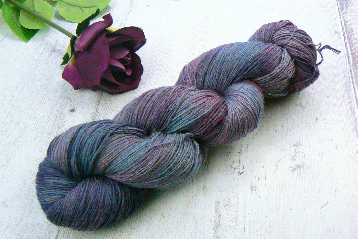 A twisted hank of variegated yarn in shades of blue, purple, and gray, styled on a white wooden surface next to a dark purple rose.