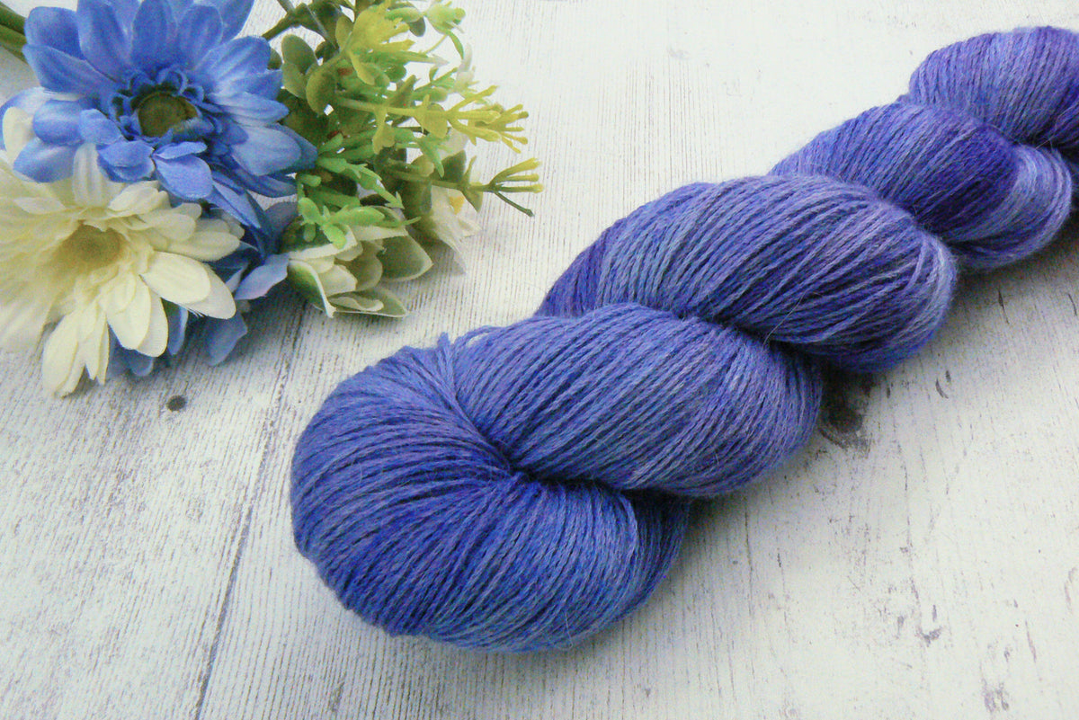 A single skein of variegated blue and purple yarn placed diagonally on a white wooden surface, styled with blue and white flowers in the upper left corner.