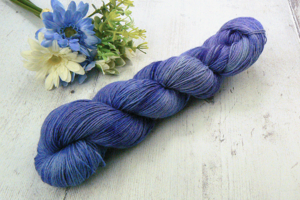 A twisted skein of variegated blue and purple yarn placed diagonally on a white wooden surface, styled with small blue and white flowers in the upper left corner.
