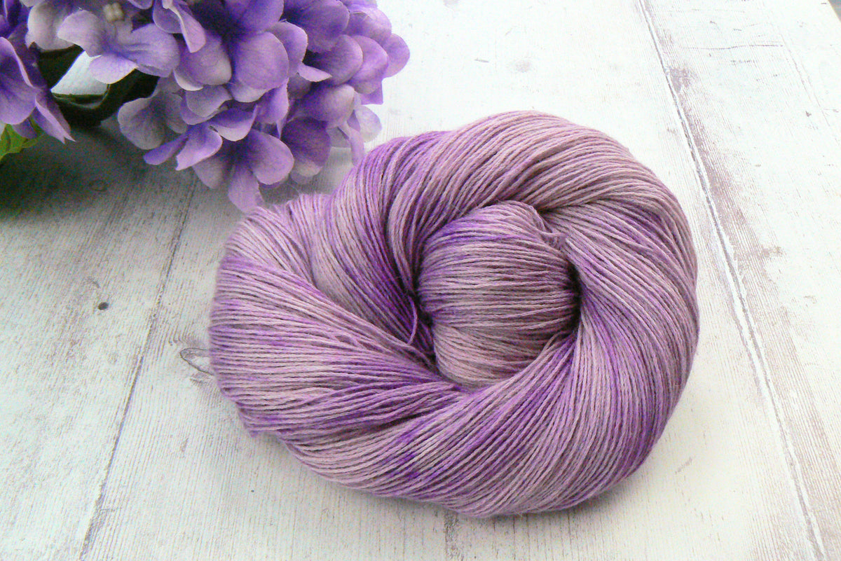 A single skein of variegated lavender and purple yarn placed on a white wooden surface, with purple flowers partially visible in the upper left corner.