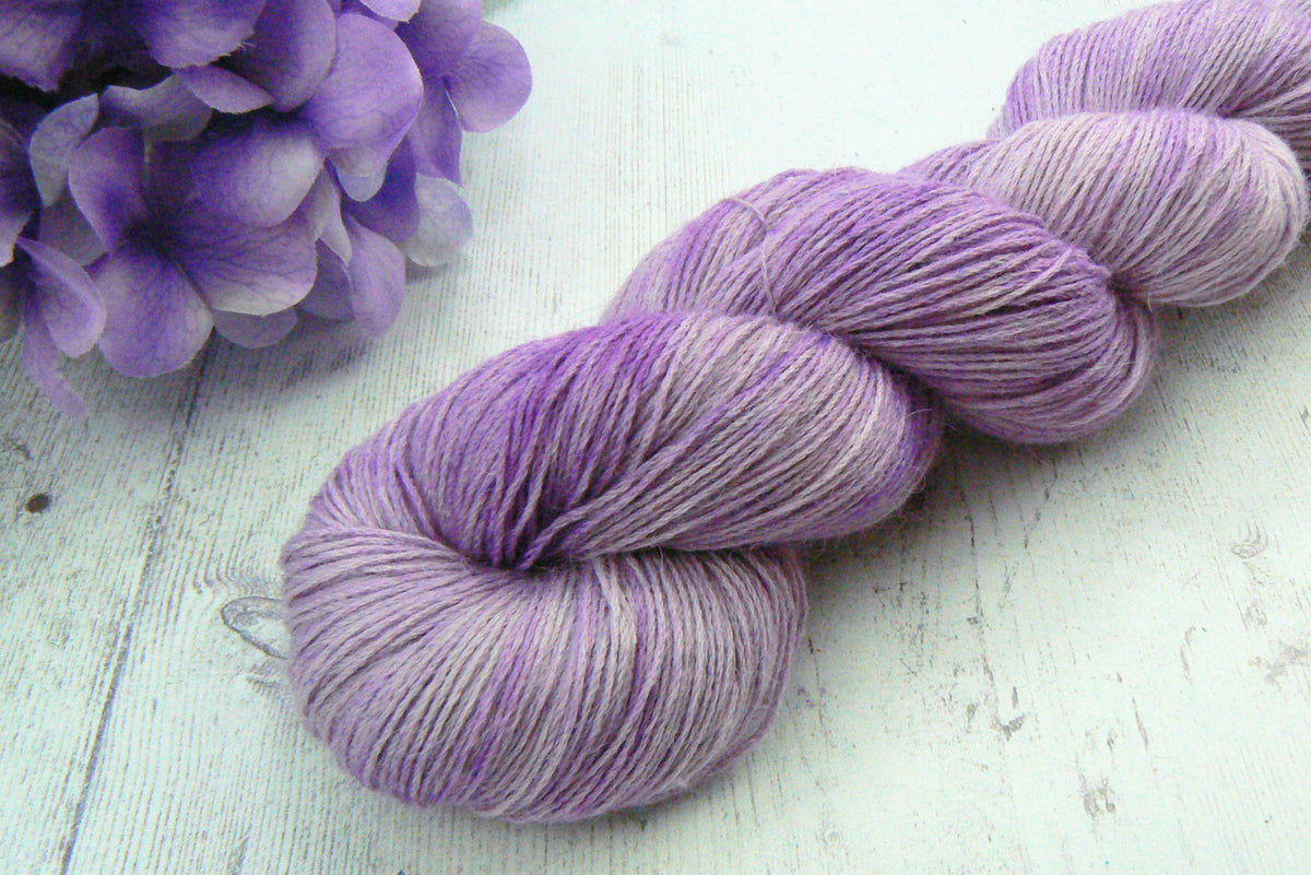 A skein of variegated lavender yarn arranged diagonally on a white wooden surface, with purple hydrangea flowers in the top-left corner.