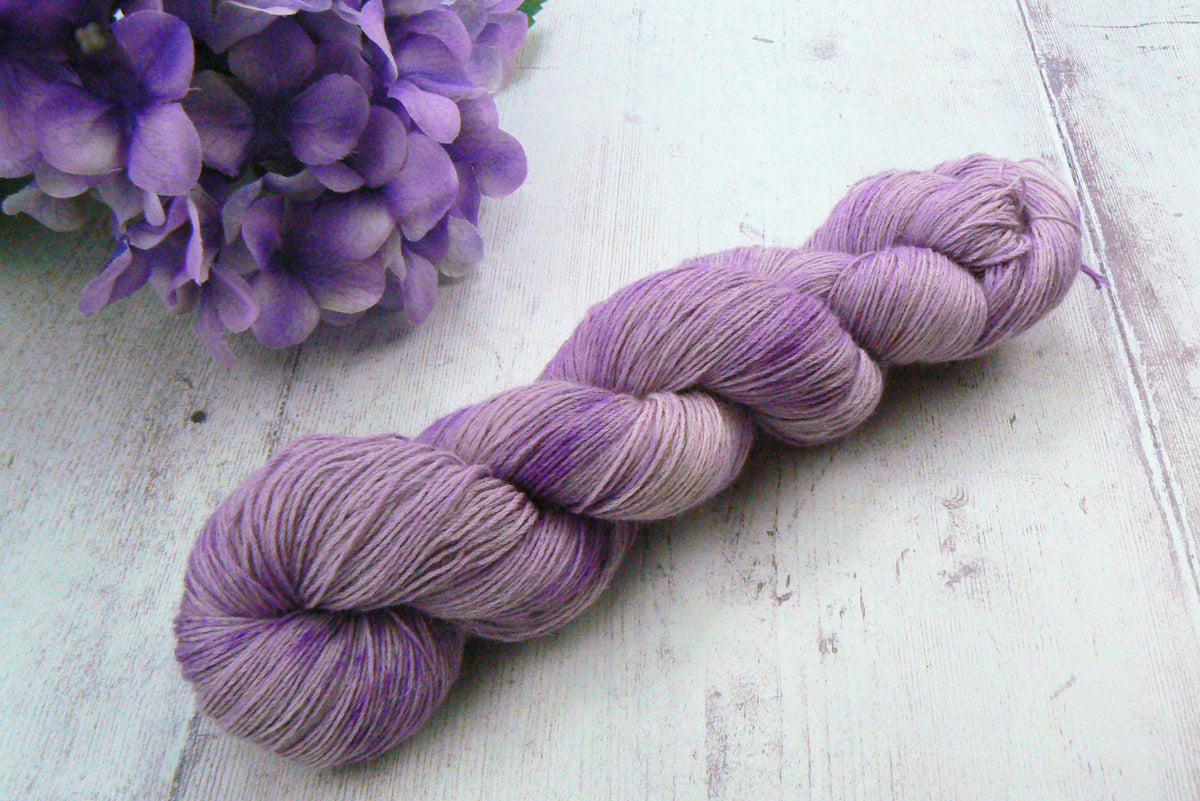 A twisted skein of variegated lavender and purple yarn displayed diagonally on a white wooden surface, with a decorative purple flower in the top-left corner.