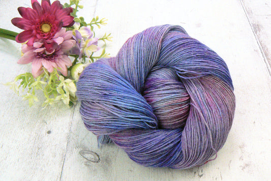 A skein of variegated yarn in shades of purple, blue, and pink, styled on a white wooden surface next to a small bouquet of pink and purple flowers.