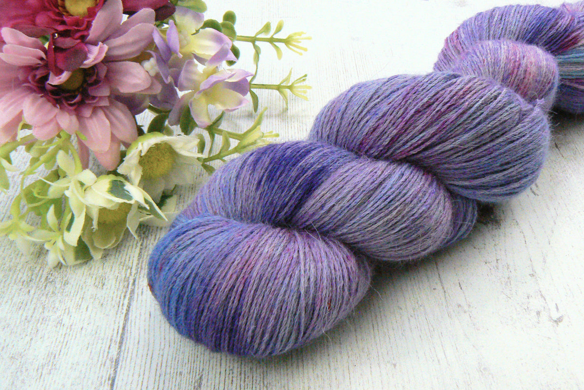 A hank of variegated purple and blue yarn styled next to pink and white flowers on a white wooden surface.