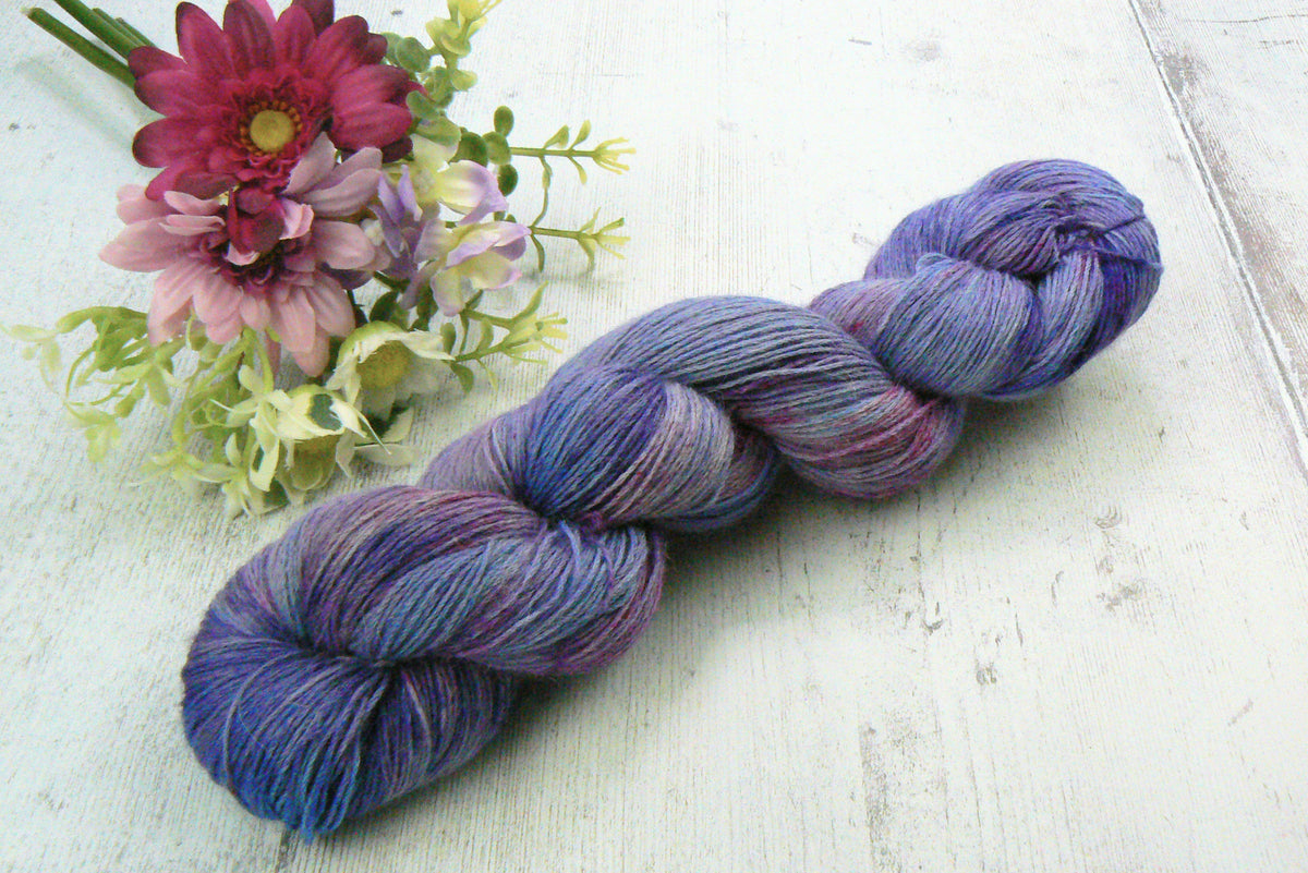 A twisted hank of variegated yarn in shades of purple, blue, and pink, styled on a white wooden surface next to a small bouquet of pink and purple flowers.