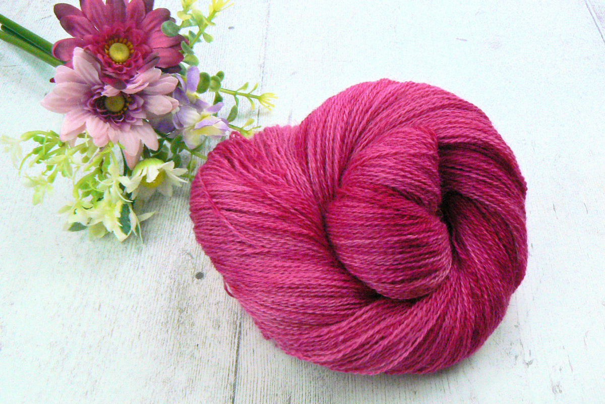 A skein of vibrant magenta pink yarn placed on a white wooden surface next to a small bouquet of pink and purple flowers.