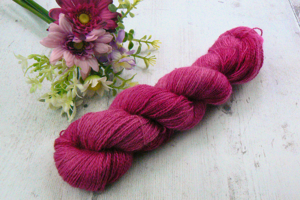 A skein of vibrant magenta pink yarn arranged diagonally on a white wooden surface next to a small bouquet of pink and purple flowers.
