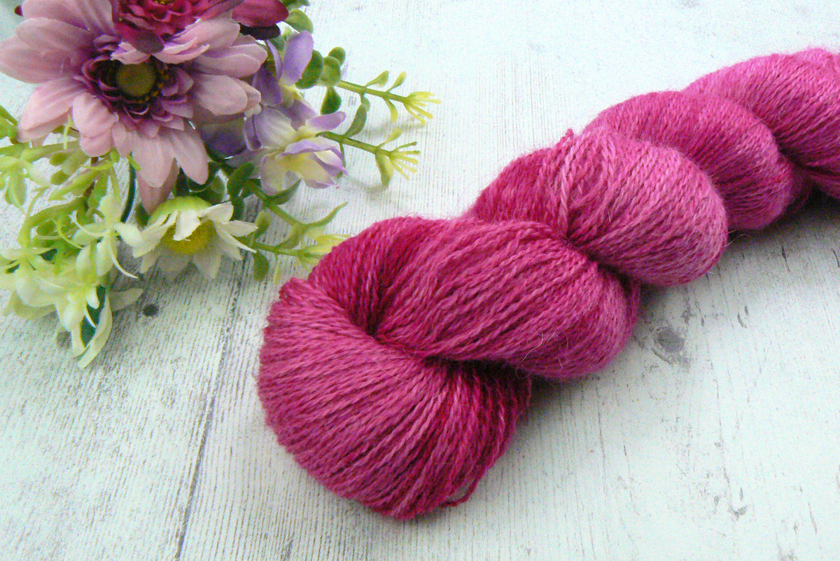A skein of bright magenta pink yarn arranged diagonally on a white wooden surface next to a decorative flower arrangement.