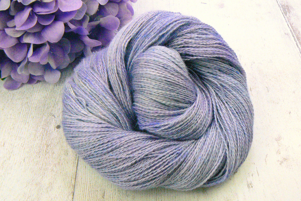 A skein of variegated lavender and light blue yarn arranged in a spiral, styled on a light wood surface next to purple hydrangea flowers.