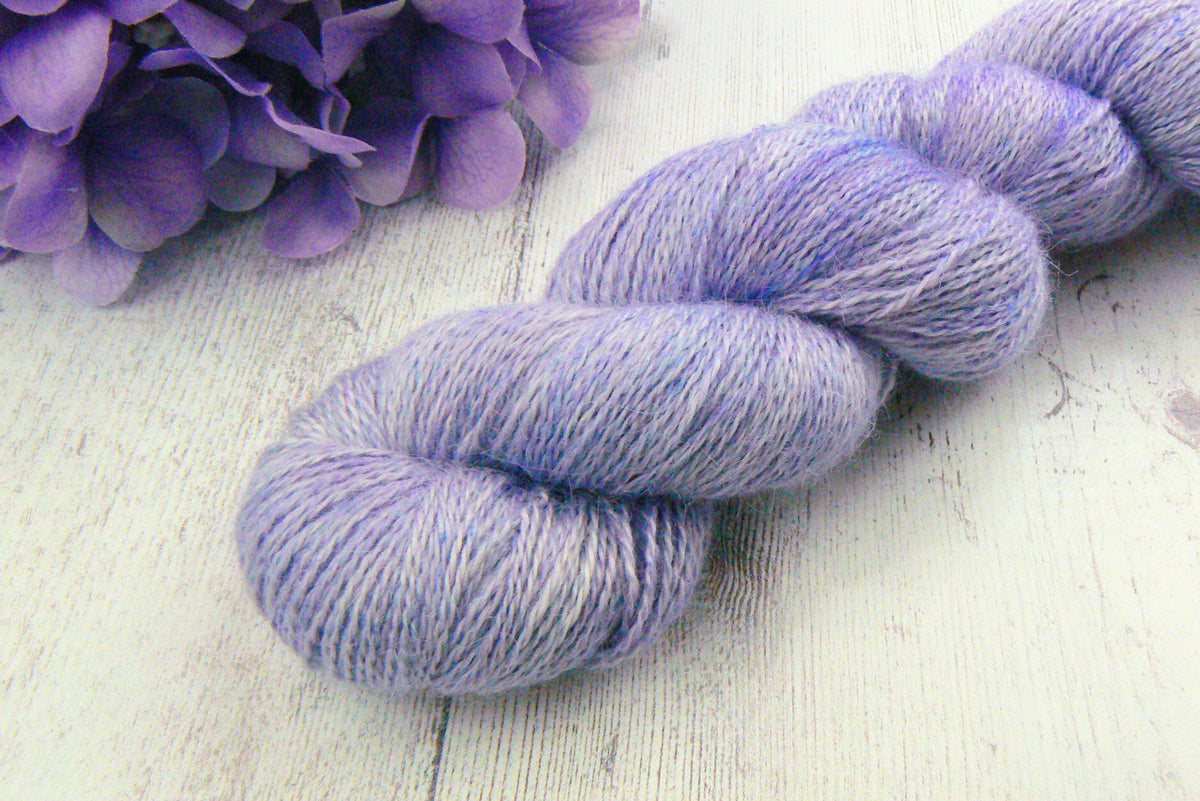 A twisted skein of lavender purple variegated yarn resting on a white wooden surface next to purple flowers.