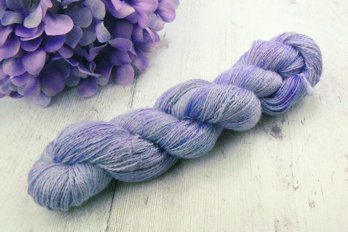 A twisted skein of lavender and periwinkle blue yarn resting on a white wooden surface next to purple hydrangea flowers.